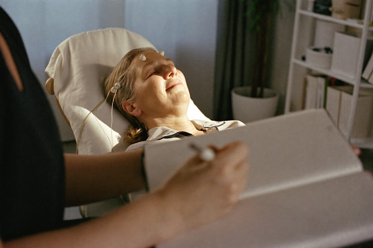 A practitioner writes in a paper notebook in the foreground while a woman rests in a reclining chair with small sensors at her temples.