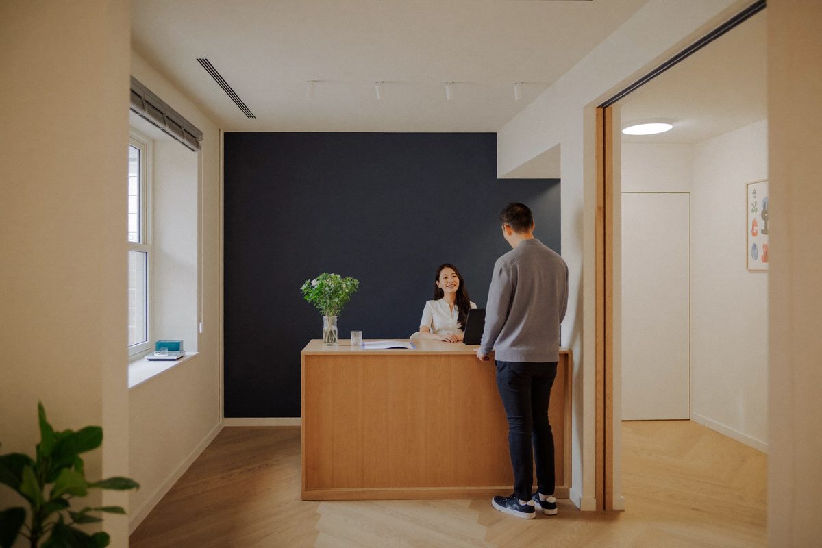 Rendered visualization of the Suite 100 reception: a light oak front desk with a receptionist seated behind it and a visitor standing at the desk, in front of a deep navy wall.