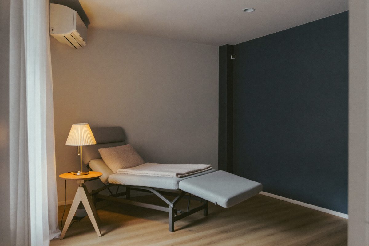 Rendered visualization of a session room with a reclining chair, a folded blanket, a small side table, and a dimmable lamp beside a curtained window.