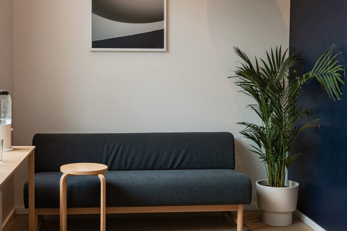 Rendered visualization of the quiet waiting area with a low upholstered bench, a small round side table, a water dispenser with glasses, and a tall plant.